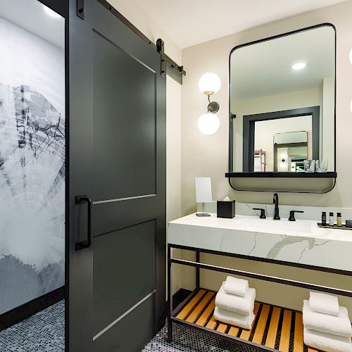A modern bathroom with a sliding door, sink, large mirror, wall lights, toiletries, flower vase, and neatly folded towels on shelves.