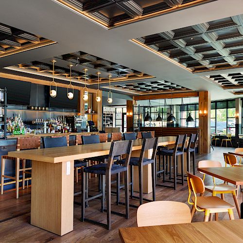 A modern bar and restaurant with wooden tables, high chairs, and a well-stocked bar counter under decorative lighting and ceilings.