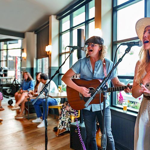 Two musicians are performing in a cozy, well-lit room while an audience is seated, attentively listening.
