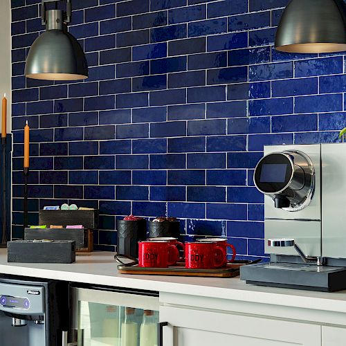 A modern kitchen with blue tile backsplash, pendant lights, a coffee machine, red canisters, candles, and potted plant on a white countertop.