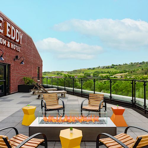 An outdoor seating area at The Eddy Taproom & Hotel with a fire pit, surrounded by mountains and greenery, under a clear sky.