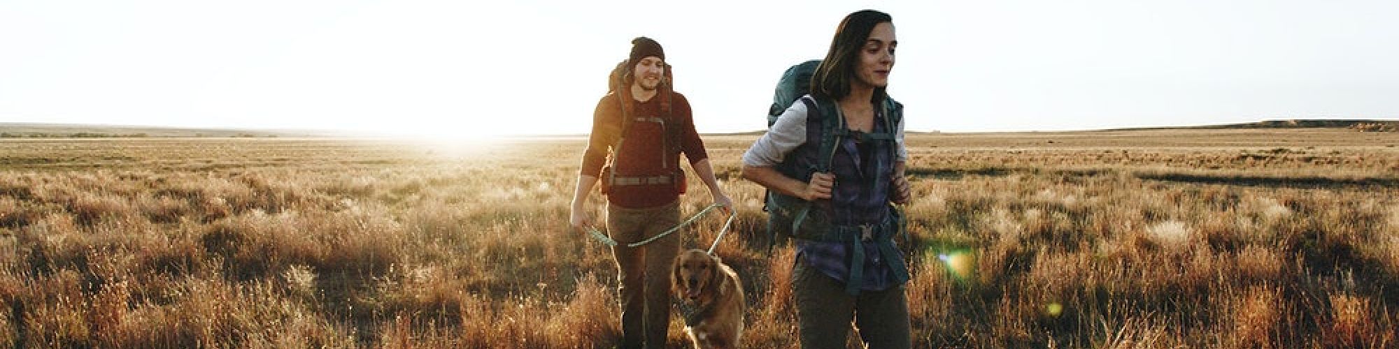Two people are hiking through a grassy field at sunrise or sunset, carrying backpacks and enjoying the outdoors.