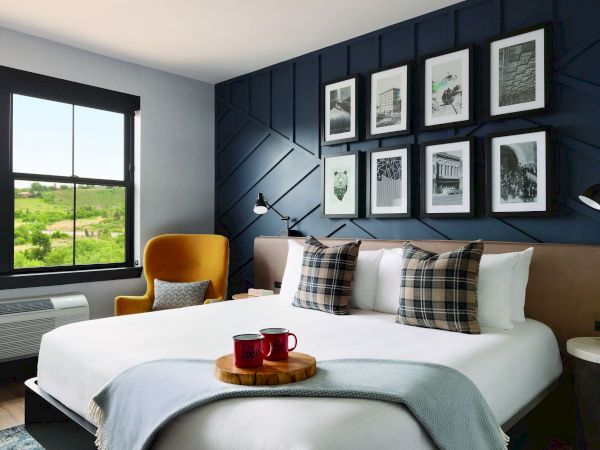 A cozy bedroom with a modern design, featuring a large bed, plaid pillows, a gallery wall, an armchair by the window, and a tray with mugs on the bed.