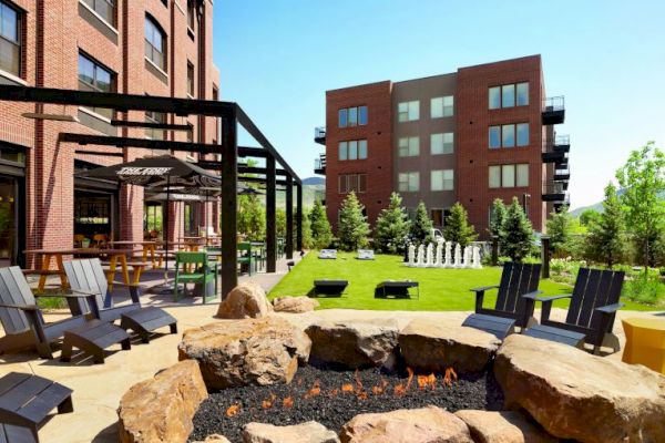 The image shows a modern outdoor area with a fire pit, Adirondack chairs, greenery, two brick buildings, picnic tables, and a giant chess set.