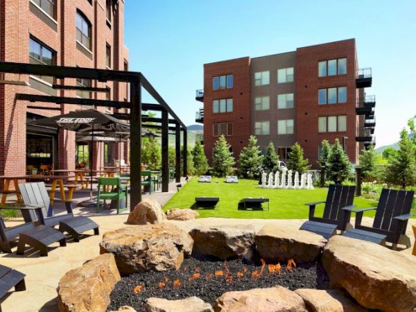 The image shows a modern outdoor area with a fire pit, Adirondack chairs, greenery, two brick buildings, picnic tables, and a giant chess set.