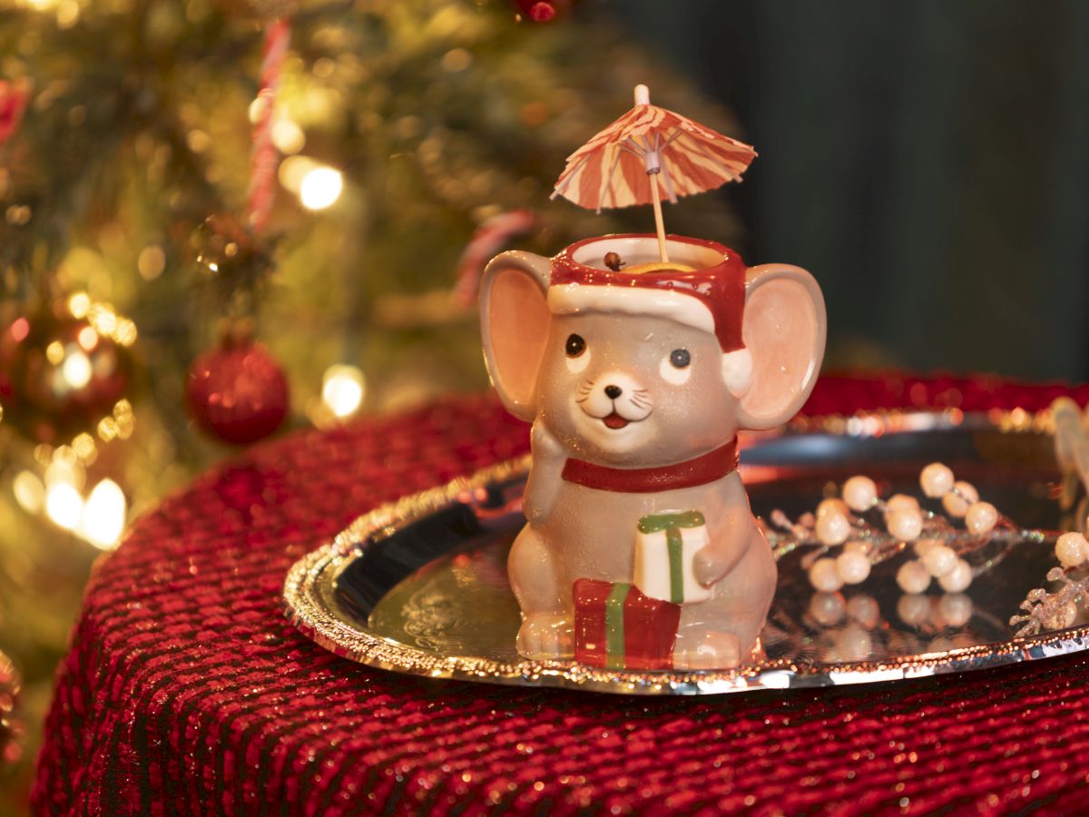 A cute ceramic mouse ornament with a tiny red-and-white umbrella sits on a Christmas scene with warm lights and beads.
