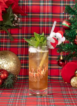 A festive holiday cocktail sits on plaid with pine, ornaments, and a cute koala in a Santa hat sipping through a straw.