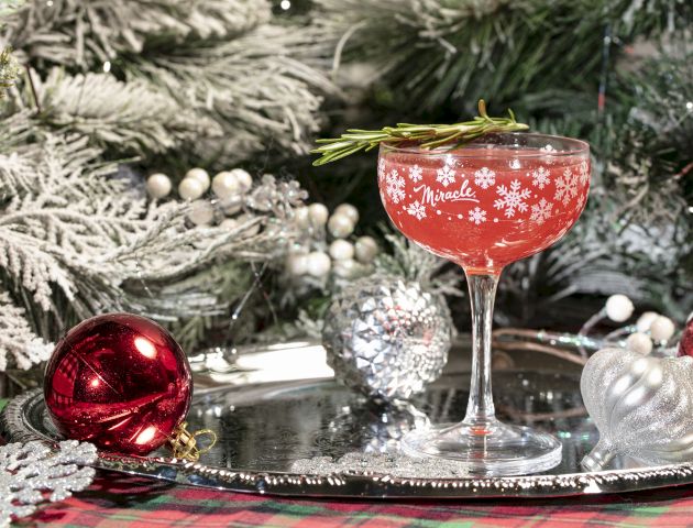 A &ldquo;Miracle&rdquo; red holiday drink in a stemmed glass sits on a festive tray with ornaments beside a frosted Christmas tree.