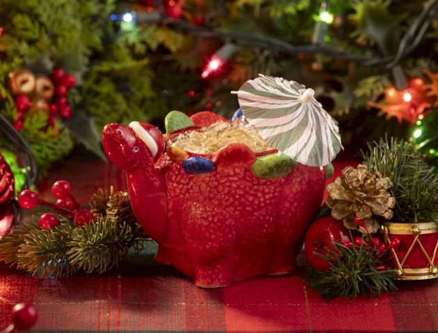 A cheerful holiday scene with a red reindeer-like ornament wearing a small umbrella, surrounded by pine, ornaments, and twinkling lights.