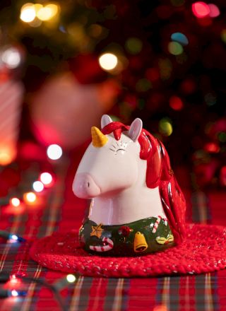 A glossy red-and-white unicorn figurine with a tiny horn sits on a red plaid surface as holiday lights blur behind it.