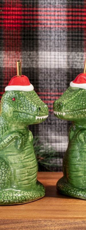 A festive Christmas display shows two green alligator-like mugs wearing Santa hats amid ornaments, gifts, and pine decorations on a plaid backdrop.