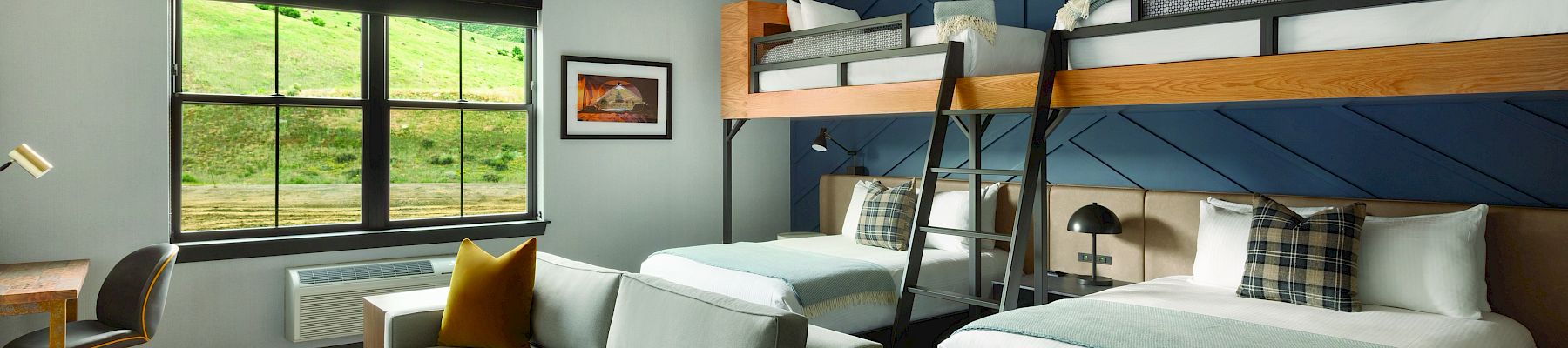 A modern room features a bunk bed, a queen bed, a sofa, a desk, and a window with a scenic view. Decor includes pillows and framed art.