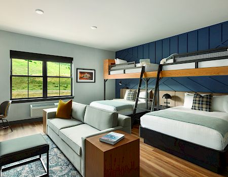 A modern room features a bunk bed, a queen bed, a sofa, a desk, and a window with a scenic view. Decor includes pillows and framed art.
