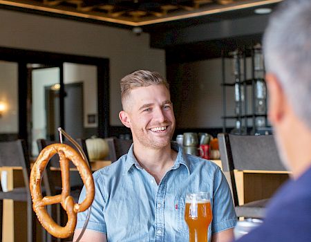 Two men are sitting at a table, one with a giant pretzel and a glass of beer, smiling in a restaurant setting.