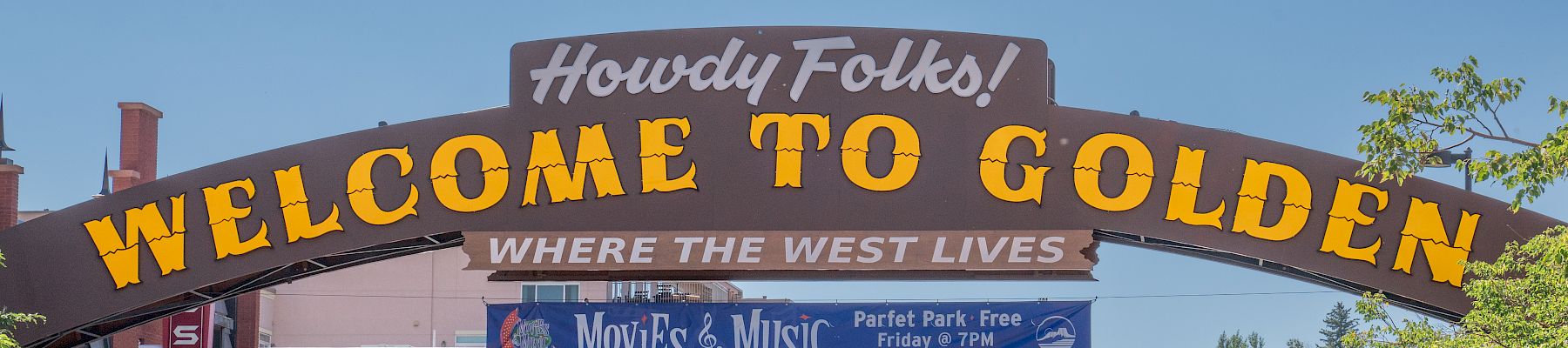 A sign reads "Welcome to Golden" on an archway over a street lined with buildings and trees under a clear blue sky.