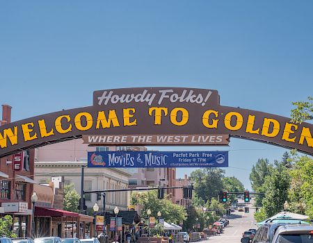 A large archway sign reads, "Howdy Folks! Welcome to Golden, Where the West Lives," above a busy street with shops and trees.