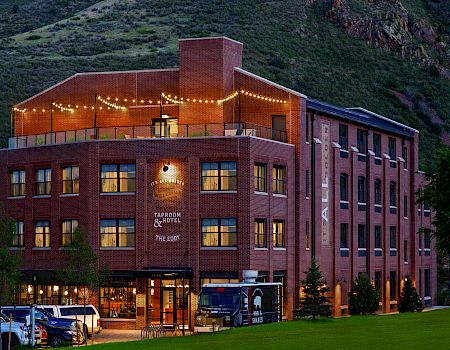 A brick building with a wrap-around porch, string lights, a sign for a tavern and hotel, and a parked bus in front of a green hillside.