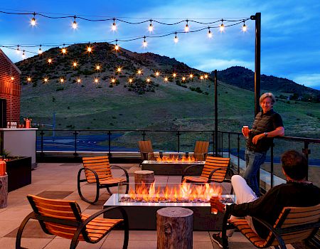Two people are relaxing by outdoor fire pits on a rooftop patio, with string lights overhead and scenic mountains in the background.