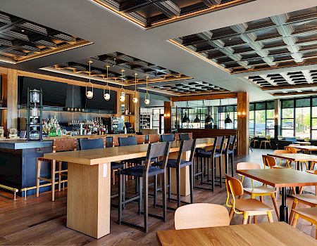 The image shows a modern bar and restaurant with high tables, chairs, and a bar counter, featuring a stylish ceiling and plenty of natural light.