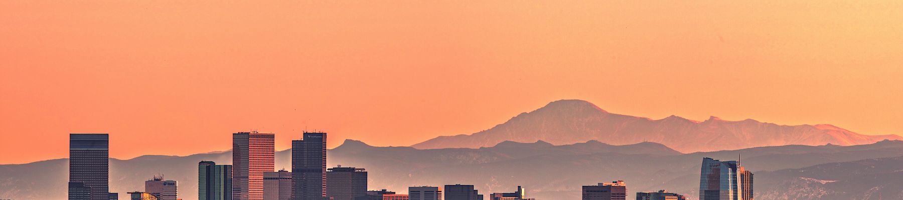 A city skyline with tall buildings at sunset, with mountains visible in the background under a clear, colorful sky.