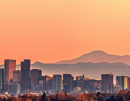 A city skyline with tall buildings at sunset, with mountains visible in the background under a clear, colorful sky.