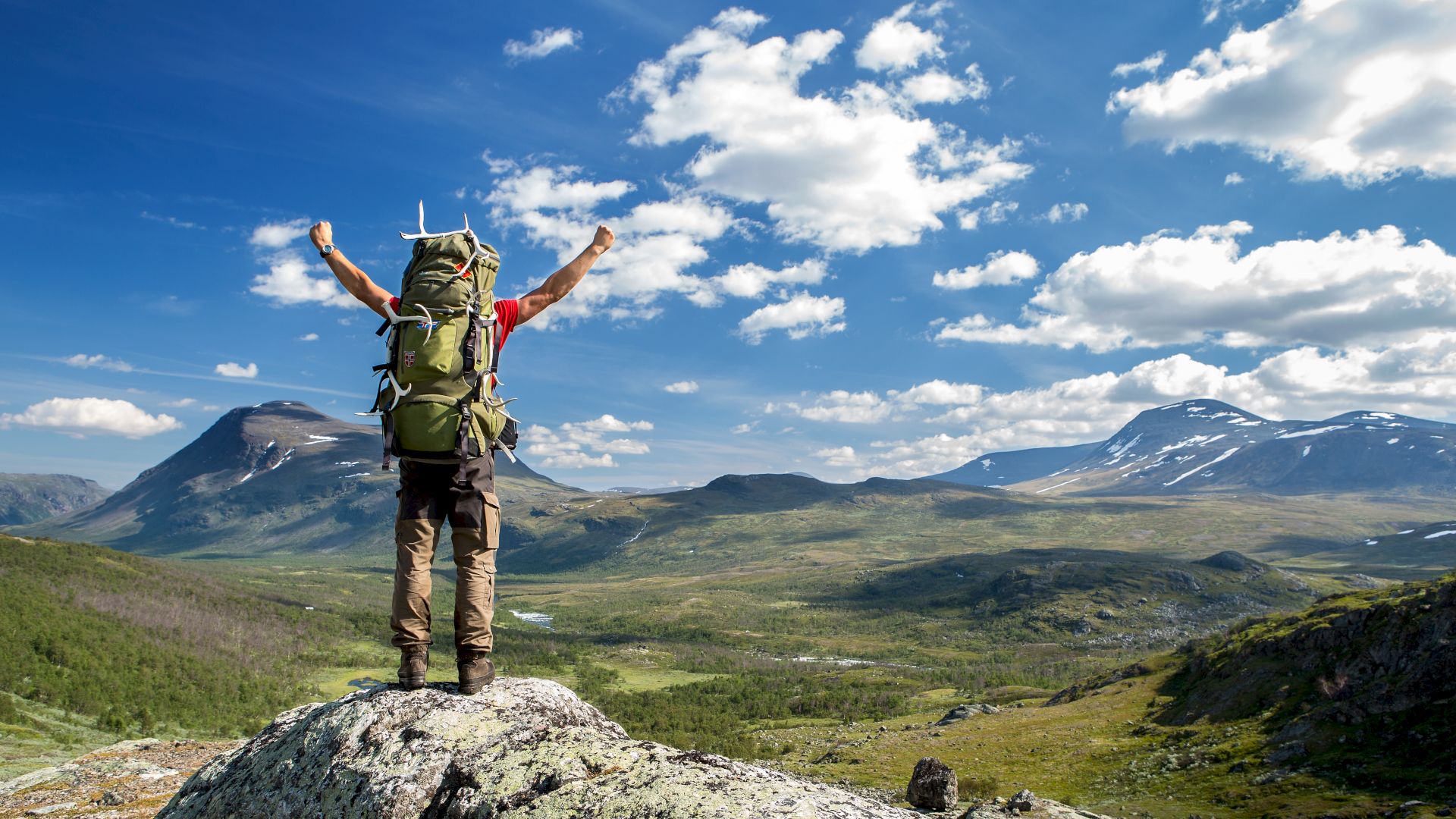 A hiker with a large backpack stands triumphantly on a rock, arms raised, overlooking a vast, scenic mountain landscape under a bright, blue sky.