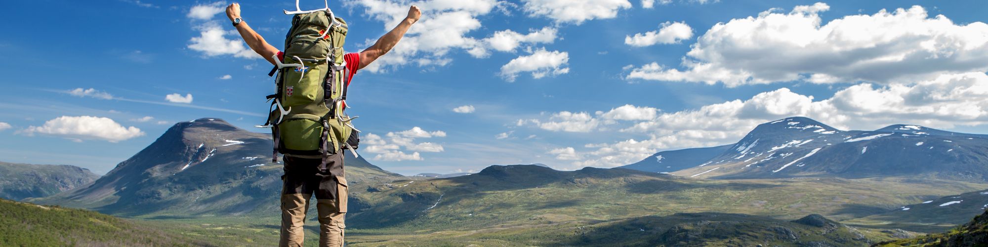 A hiker with a large backpack stands triumphantly on a rock, arms raised, overlooking a vast, scenic mountain landscape under a bright, blue sky.