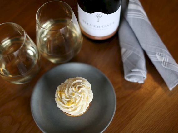 Two glasses of white wine, a bottle, a small plate with a swirl-topped cupcake or frosting, and a folded napkin on a wooden table.