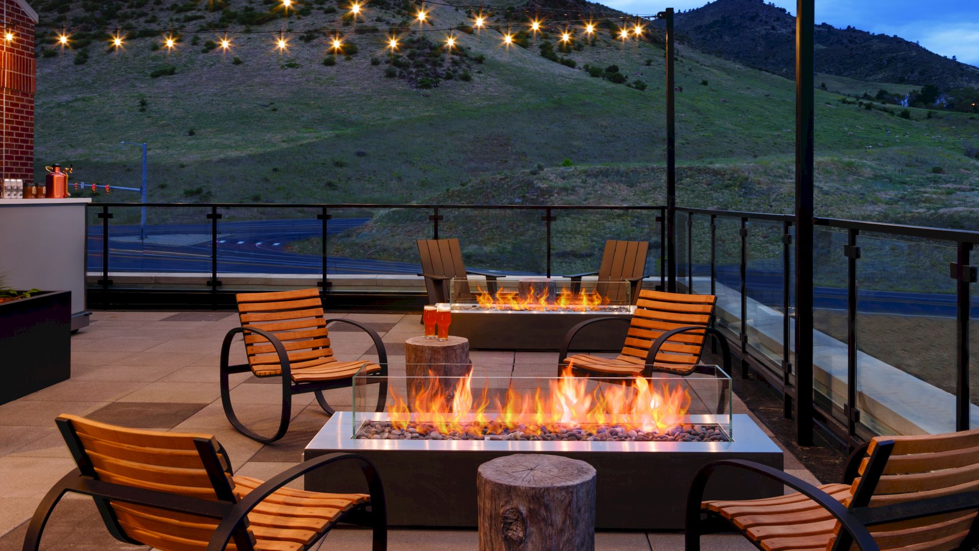 A cozy outdoor patio with several wooden chairs, fire pits, hanging string lights, and scenic hills in the background at dusk, ending the sentence.