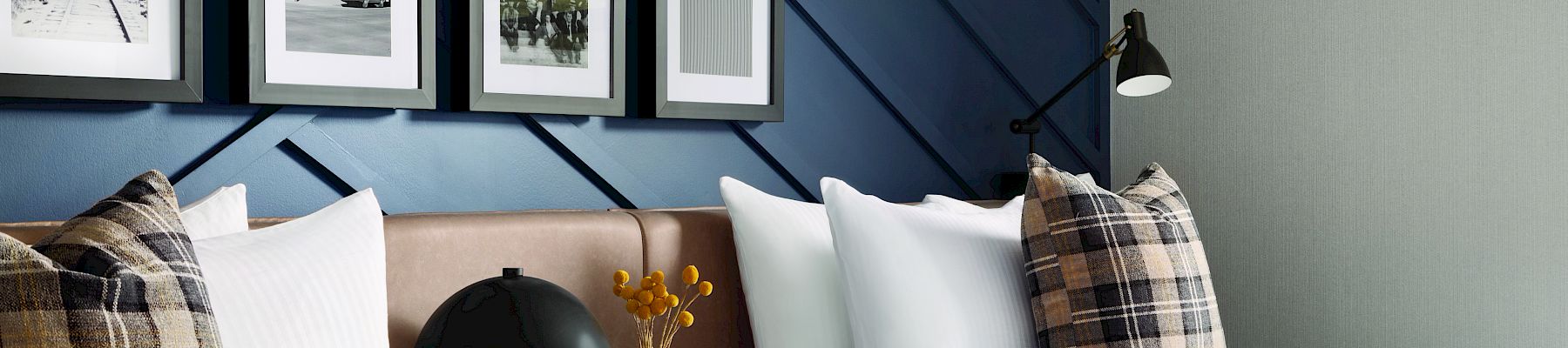 A modern bedroom features two beds with plaid pillows, a gallery wall of photos, and a black lamp on a shared nightstand, offering a cozy vibe.
