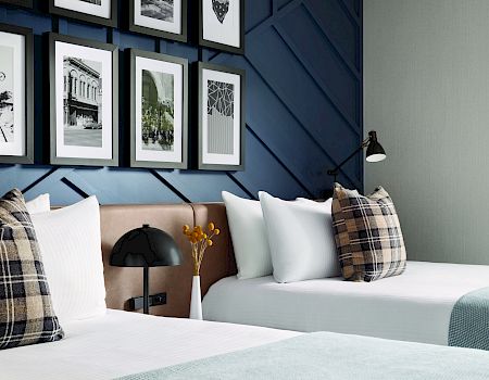 A modern bedroom features two beds with plaid pillows, a gallery wall of photos, and a black lamp on a shared nightstand, offering a cozy vibe.