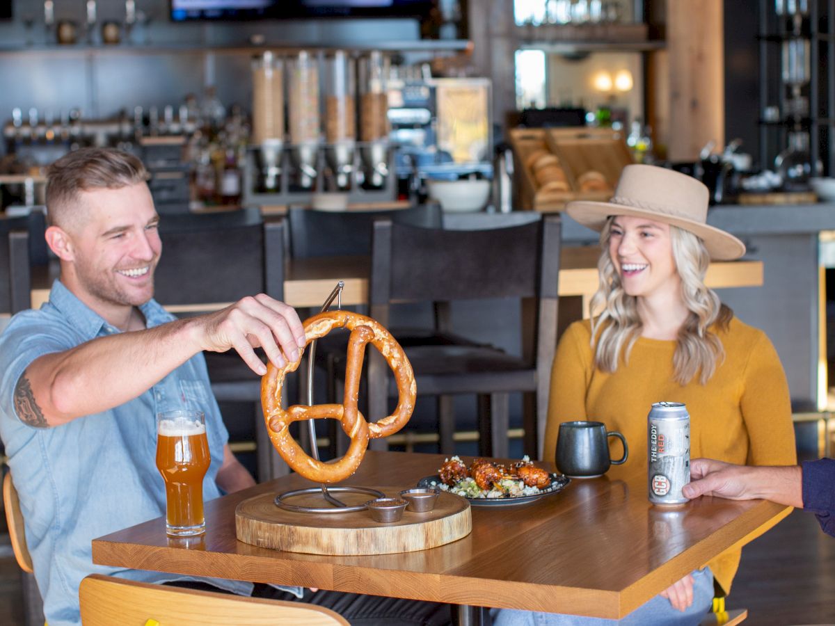 Two people are seated at a table in a modern bar, enjoying drinks and a large pretzel.
