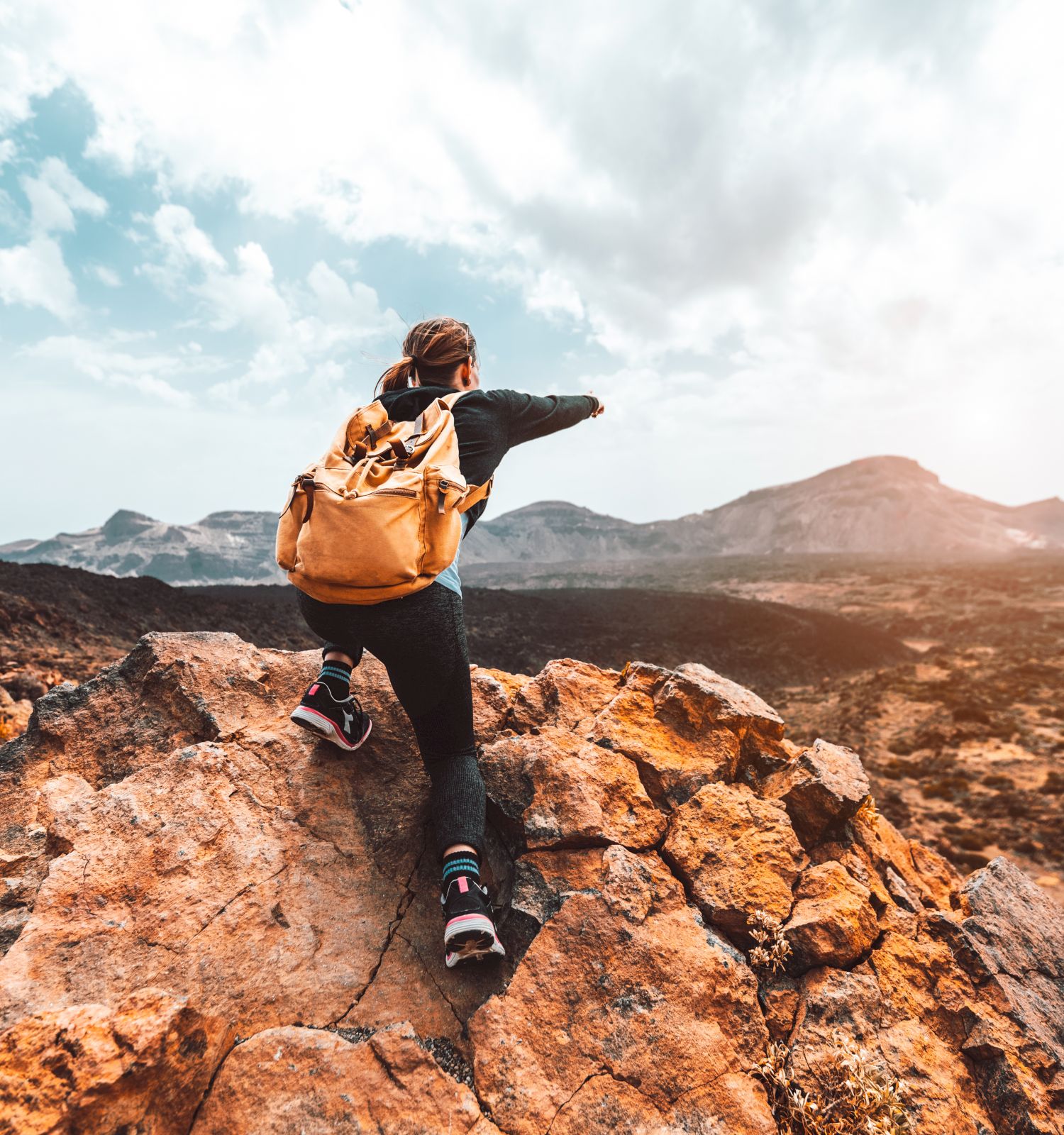 A person with a yellow backpack climbs rocky terrain, overlooking a vast landscape with distant mountains under a partially cloudy sky.