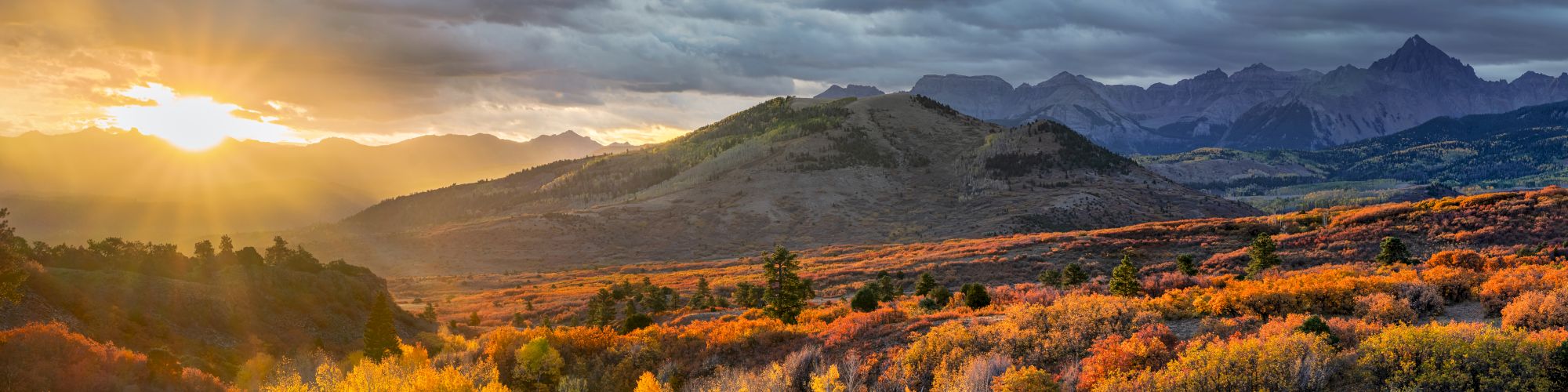 A stunning landscape with a colorful autumn forest, distant mountains, and a dramatic sunset sky.