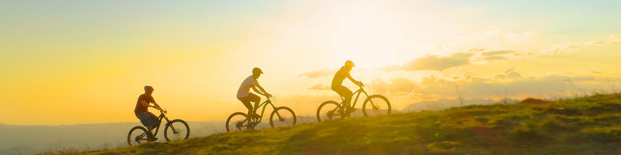 Three people are biking uphill on a grassy path during sunset, with mountains in the background.