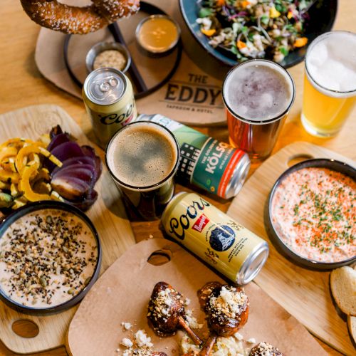Assorted appetizers and drinks, including a large pretzel, beer cans and glasses, dips, and a salad, are displayed on a wooden table.