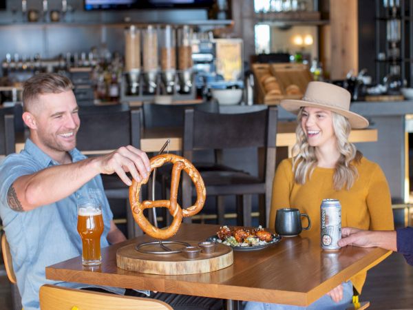 Two people enjoy pretzels and beer at a cozy cafe; a wooden pretzel stand sits between them as they chat.