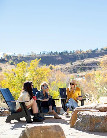 Three people are sitting outdoors on chairs, chatting near large rocks, with a scenic mountain background and autumn foliage.