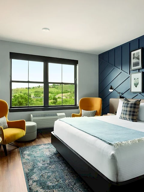 A modern hotel room with a large bed, armchairs, desk, lamp, and wall art, featuring a window with a scenic view.