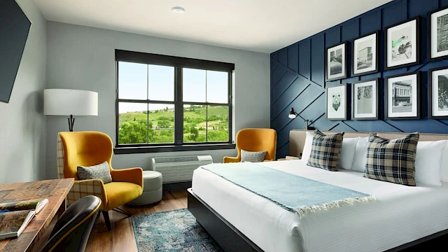 A modern hotel room with a large bed, armchairs, desk, lamp, and wall art, featuring a window with a scenic view.