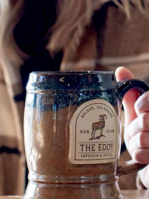 A person in a plaid shirt holds a ceramic mug labeled "The Eddy" at a wooden table, alongside a can of beverage.