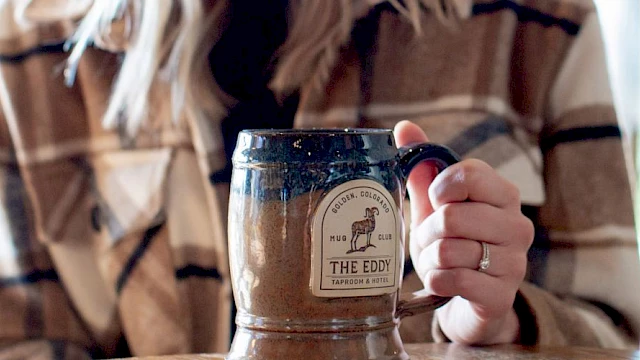 A person in a plaid shirt holds a ceramic mug labeled "The Eddy" at a wooden table, alongside a can of beverage.