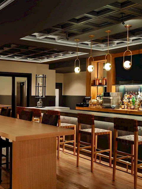 The image shows a modern bar interior with wooden stools, a long counter, and shelves stocked with various bottles.