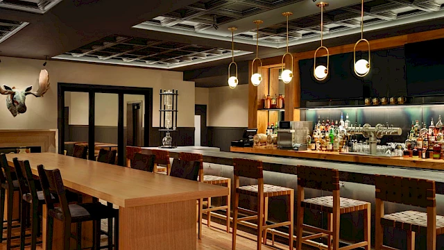 The image shows a modern bar interior with wooden stools, a long counter, and shelves stocked with various bottles.