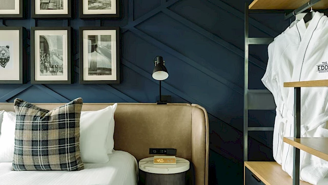 A stylish bedroom features framed photos, a plaid pillow, a nightstand with a lamp, and hanging robes on a wooden rack.
