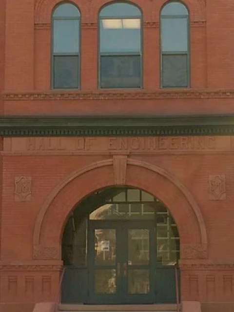 A historic brick building with arched windows and an entrance, surrounded by trees on a sunny day.