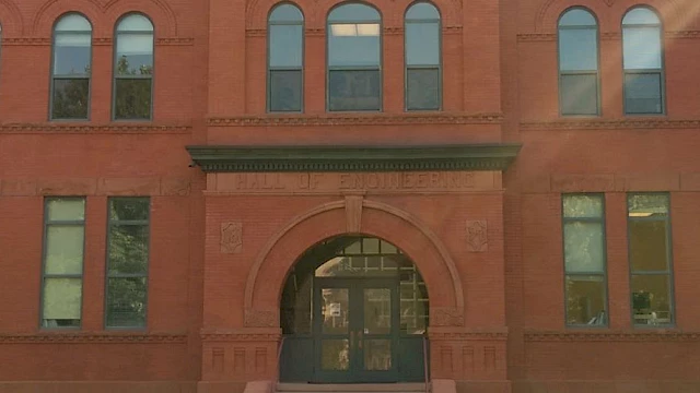 A historic brick building with arched windows and an entrance, surrounded by trees on a sunny day.