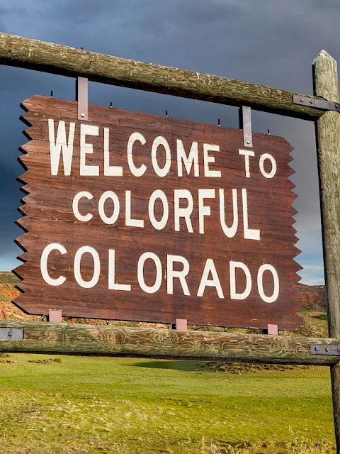 A wooden sign reads "WELCOME TO COLORFUL COLORADO," with rocky hills and green fields under a dramatic sky in the background.