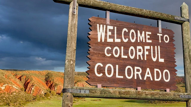 A wooden sign reads "WELCOME TO COLORFUL COLORADO," with rocky hills and green fields under a dramatic sky in the background.