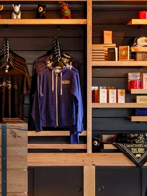 The image shows a cozy retail space with wooden shelves displaying clothing, mugs, and various items, alongside stylish wall lighting.
